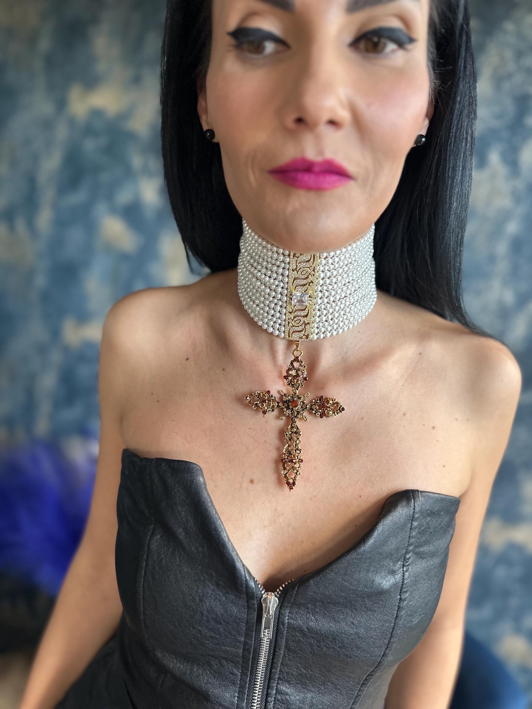 May include: A pearl choker necklace with a gold and crystal accent, paired with a decorative cross pendant. The cross is adorned with red and brown gemstones. The model is wearing a black leather top.