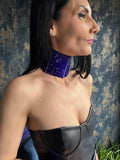 May include: A wide, blue beaded choker necklace with a gold trim. The choker is made of small, dark blue beads and is worn around the neck. The model is wearing a black leather bustier.